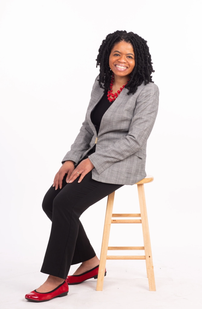 A woman in a gray blazer with black pants and red shoes sitting on a wooden stool 
